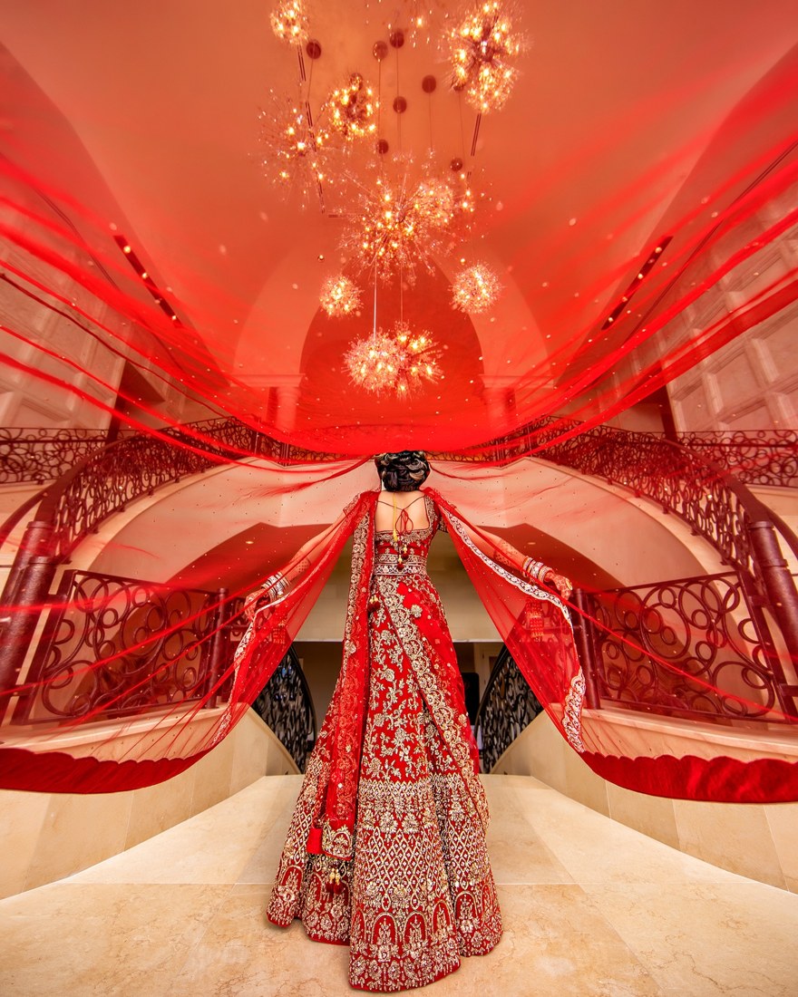 A bride beneath a flowing red veil in a chandelier-lit room